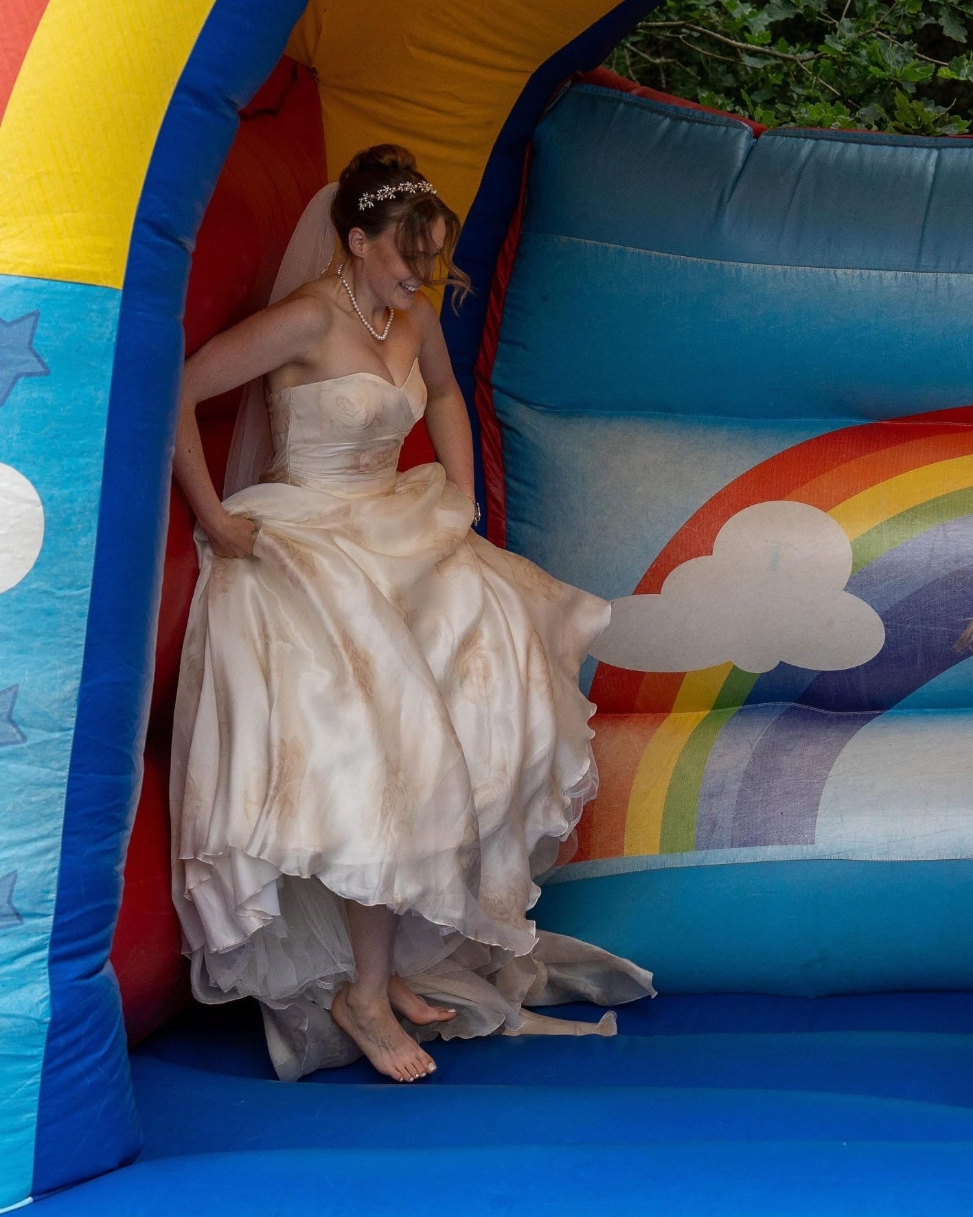 A bride in a strapless ivory satin wedding gown and veil slides down a colorful inflatable bouncy castle with rainbow and cloud graphics.